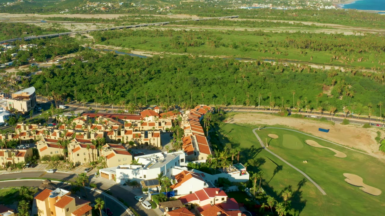 Drone view of homes, palm trees, golf fairway, and open green land.