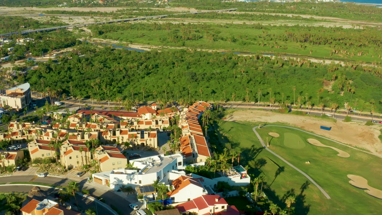 Aerial San Jose Del Cabo frame showing red-roof buildings along the lower left, a golf green and sand traps on the right, broad green fields behind them, and no visible people.