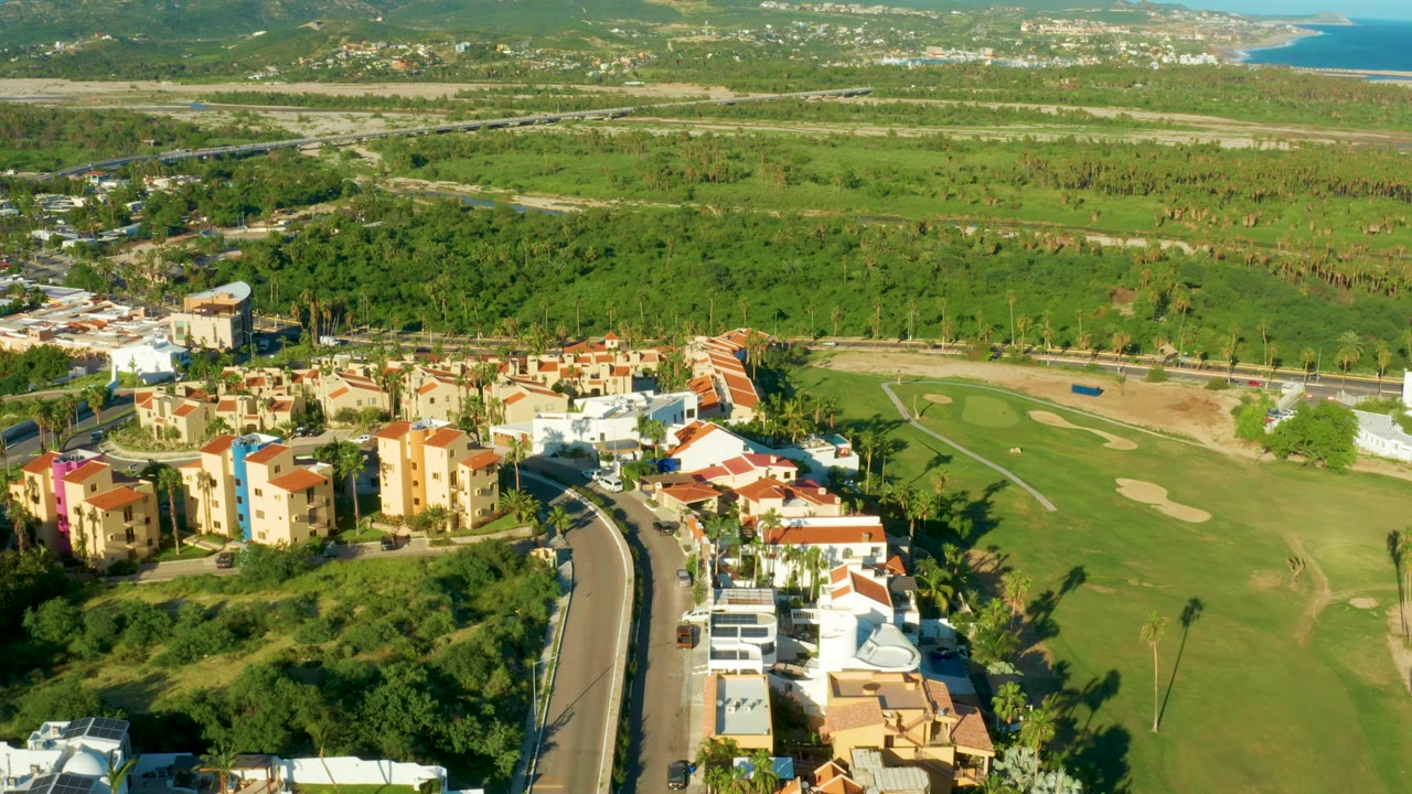 High aerial frame over San Jose Del Cabo residences, a curved road, green fairways, sand traps, distant hills, and a small ocean view, with no visible people.