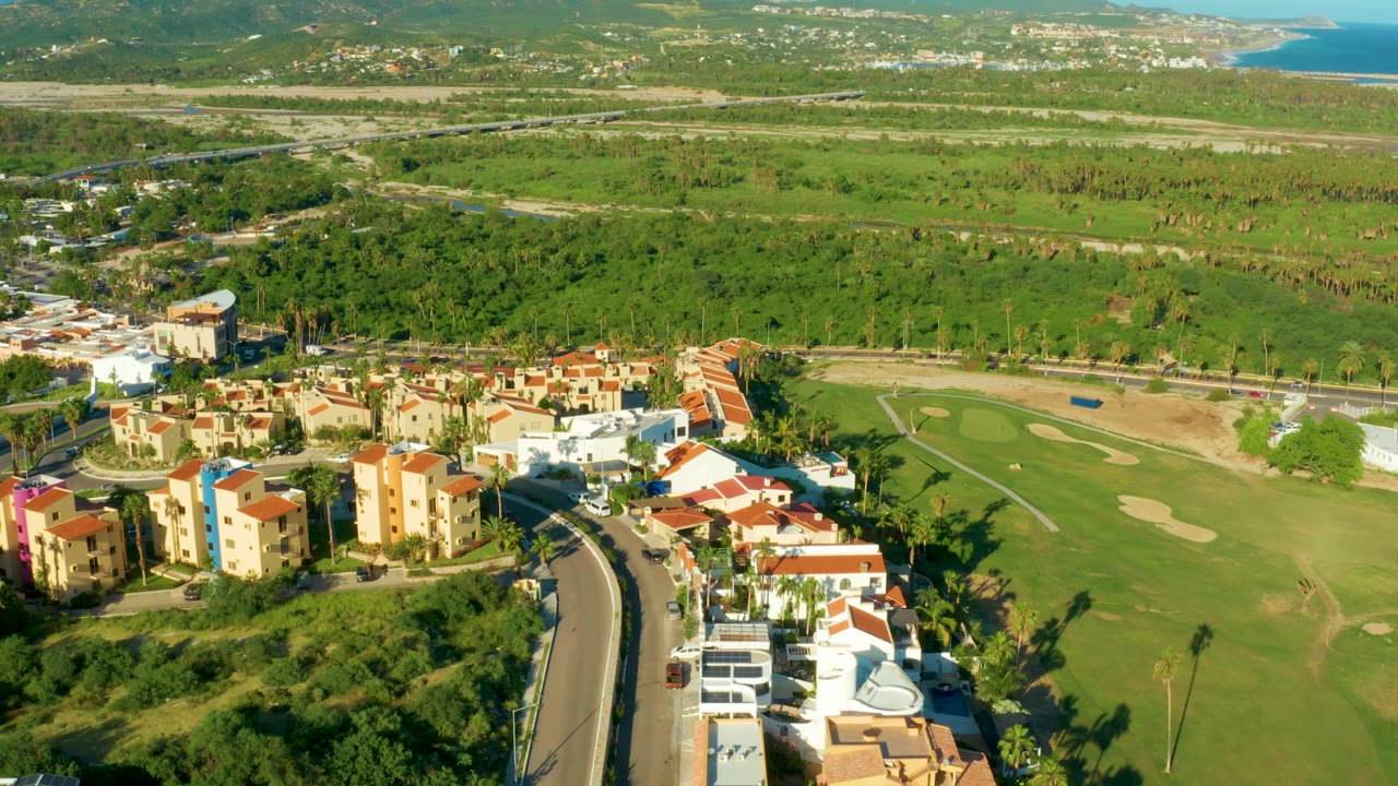 San Jose Del Cabo aerial frame with apartment-style buildings at lower left, a fairway and bunkers to the right, a broad green valley, distant hills, and ocean at the upper right, with no visible people.
