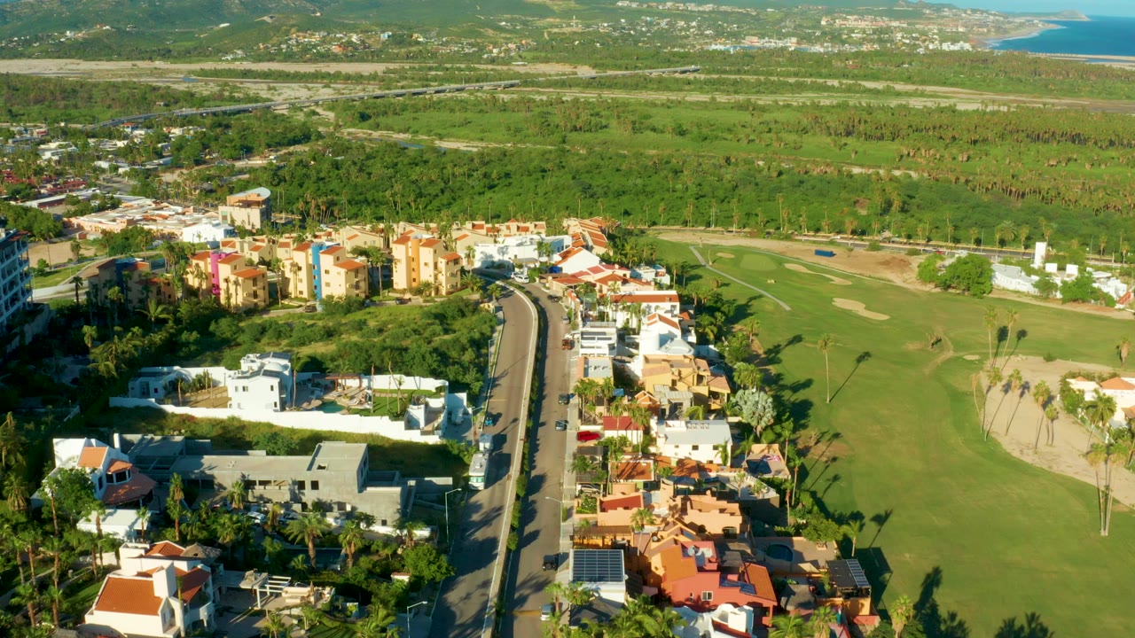Aerial frame over San Jose Del Cabo showing a curving road between white homes, colorful residential buildings, green golf fairways, palms, distant hills, a blue coastal edge, and no visible people.