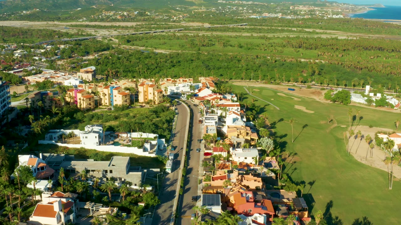 San Jose Del Cabo aerial view with the road and nearby homes slightly shifted in frame, colorful apartment-style buildings near the golf course, distant hills, coastline, and no visible people.