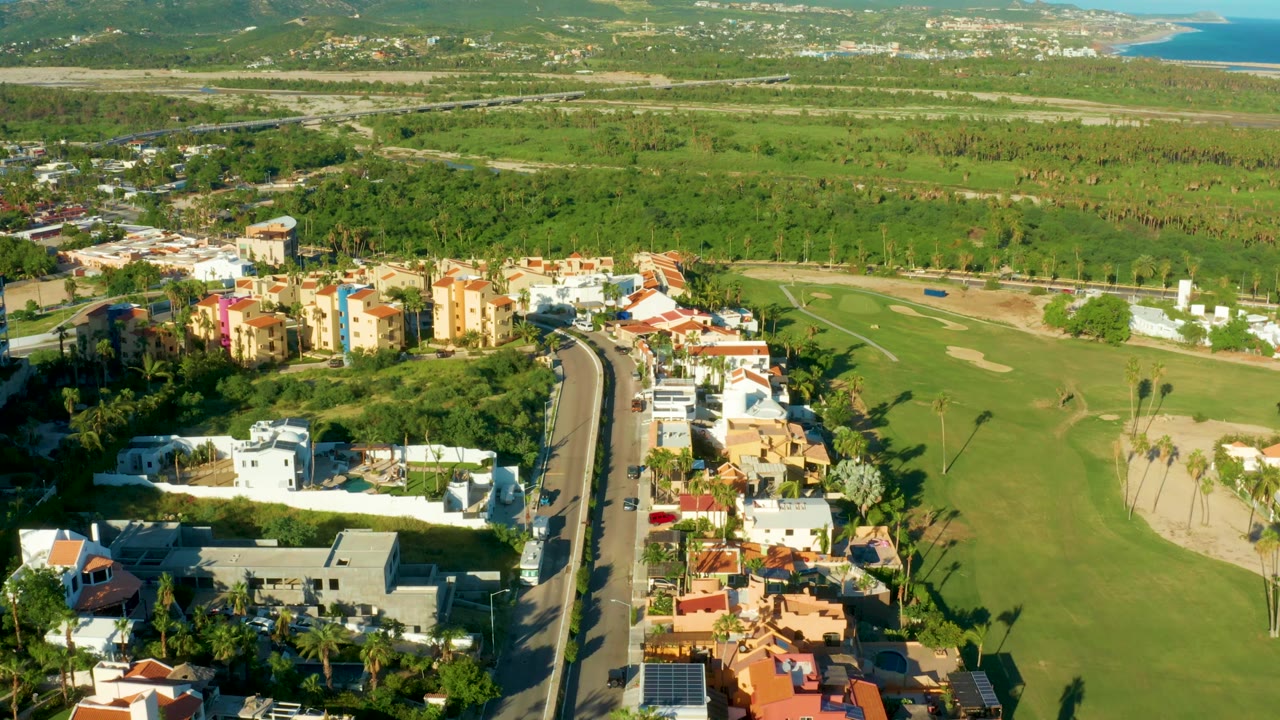 Drone frame over San Jose Del Cabo with colorful residential blocks closer to center, a road lined by homes, golf-course grass and bunkers to the right, hills in back, coastline, and no visible people.