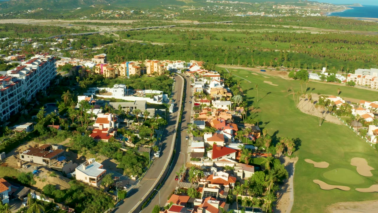 Aerial frame from San Jose del Cabo showing a curving road, red-roofed homes, a green golf fairway, sand traps, hillside scrubland, distant coastline, and no visible people.