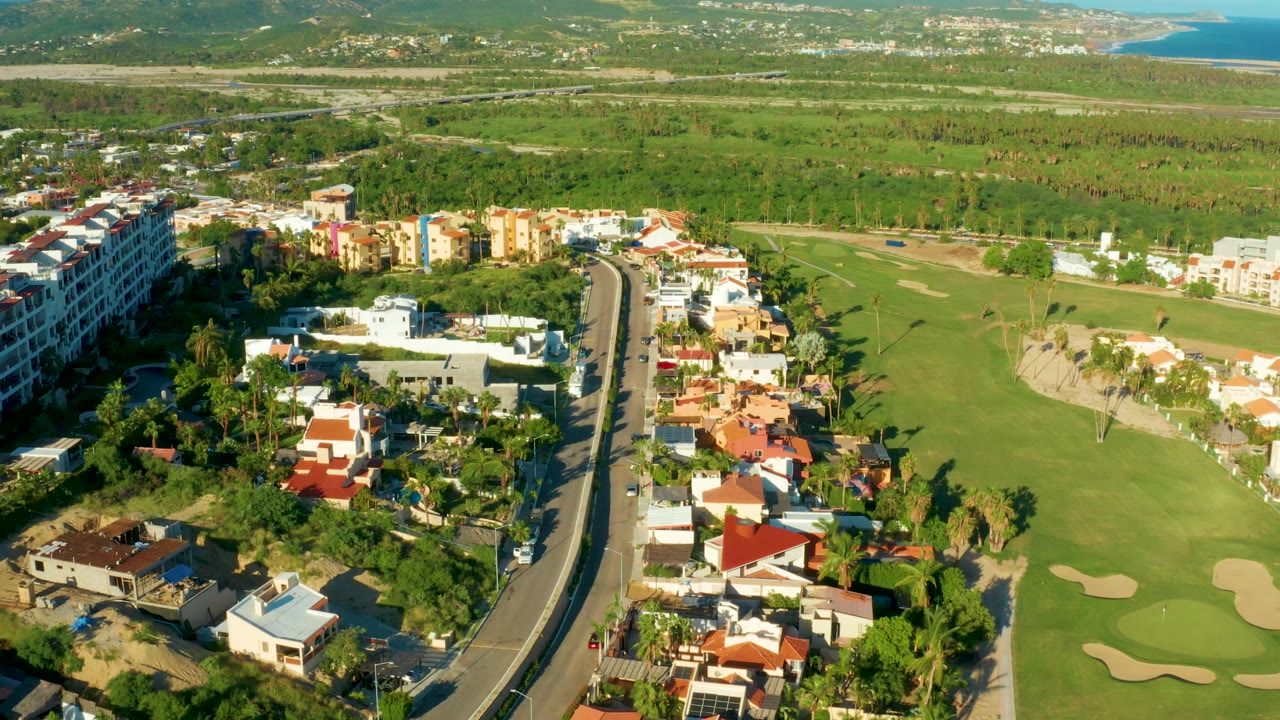 High aerial frame in San Jose del Cabo showing villas and apartments near a paved road, wide golf course grass, several sand traps, green hills, and no visible people.