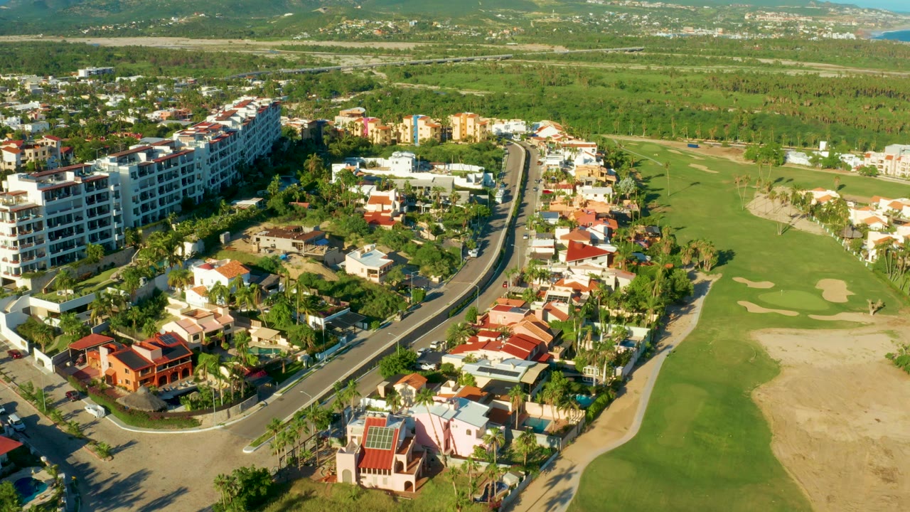 Aerial frame over San Jose Del Cabo showing a curving road between red-roof homes, multi-story buildings, and green fairways, with no visible people.