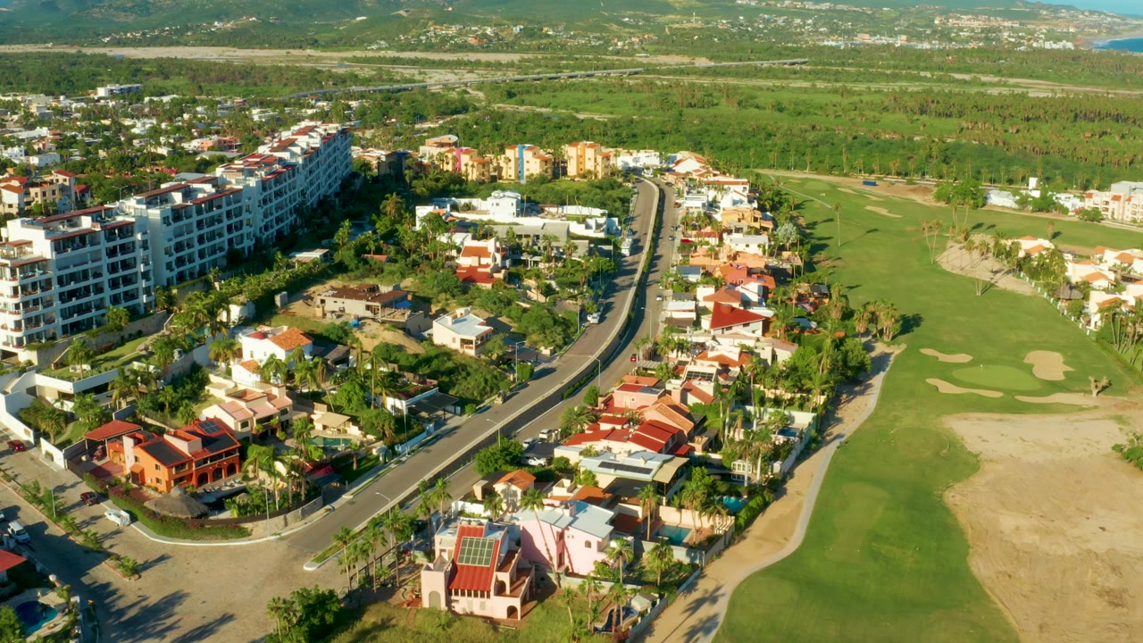Aerial frame in San Jose Del Cabo with the road centered lower in frame, white apartment-style buildings on the left, houses in the middle, and golf greens to the right, with no visible people.