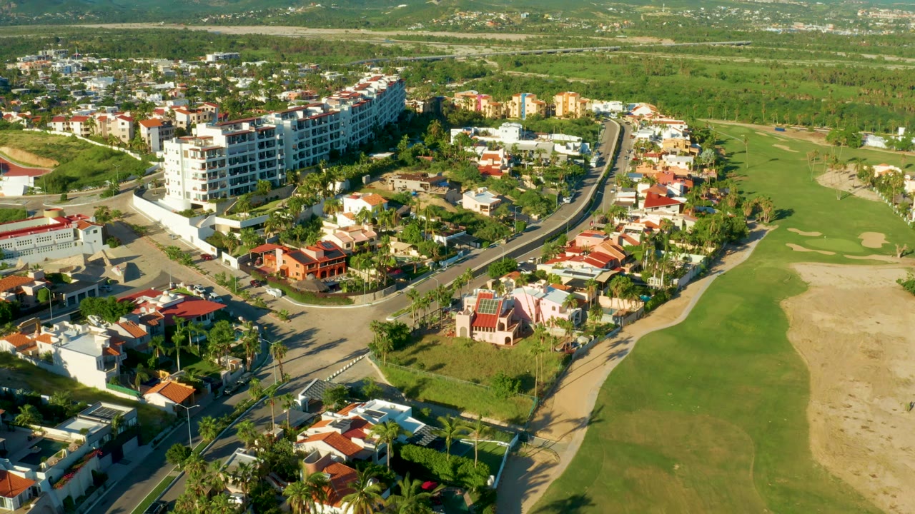 Mid-pan aerial frame over San Jose del Cabo with the curved neighborhood road centered, red and white roofs below, fairway to the right, distant hills, and no visible people.