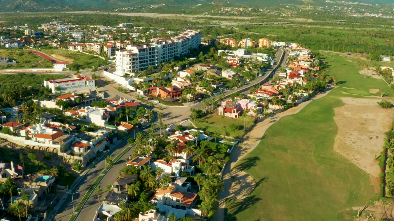 Aerial frame over San Jose del Cabo showing white and red-roofed homes beside a curved road, a green golf course fairway, distant low hills, and no visible people.