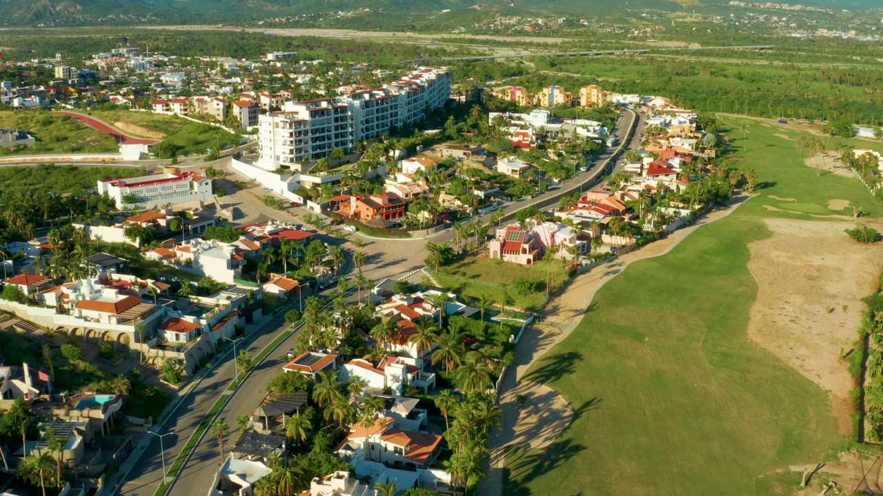 San Jose del Cabo aerial view with the curved road running through houses and palms, a large green fairway to the right, low buildings beyond, and no visible people.