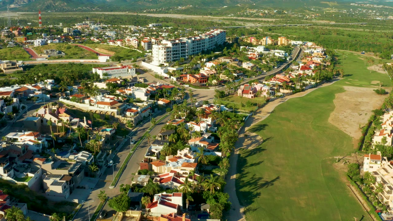 Aerial view over San Jose del Cabo homes and curving residential roads beside a broad green field, with distant hills and no visible people.