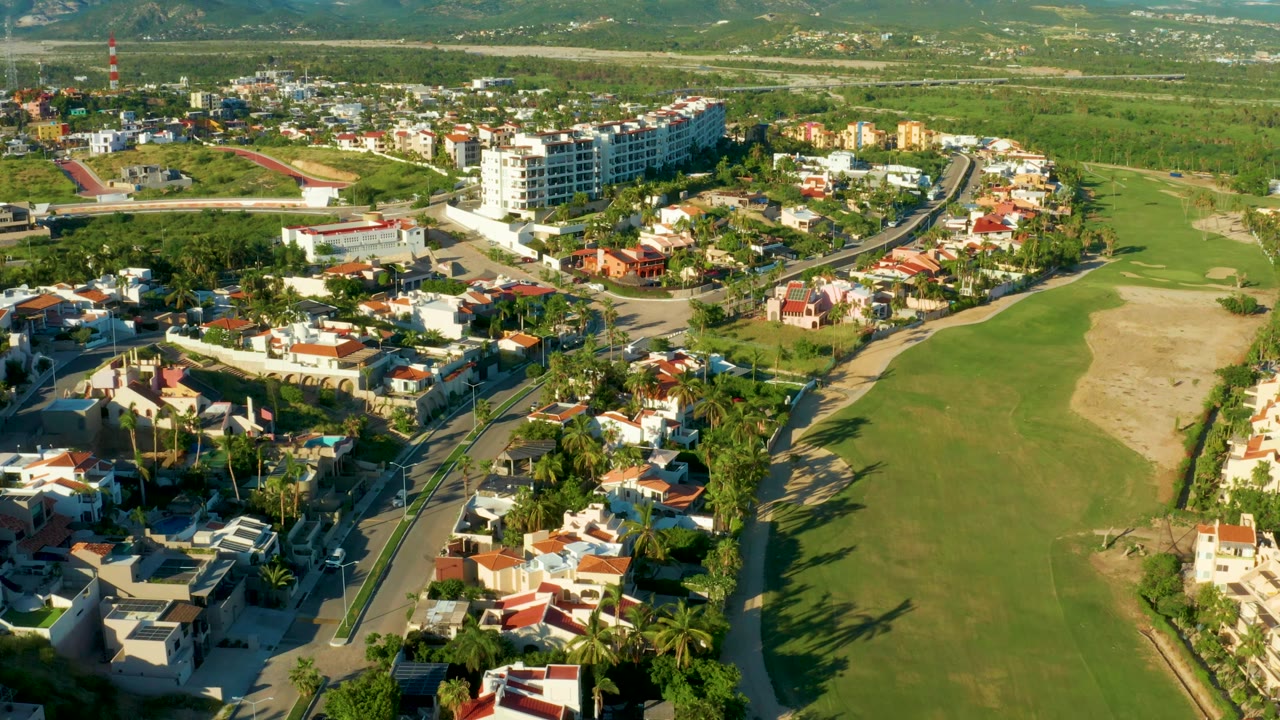 Drone-height view across San Jose del Cabo homes, palms, paved streets, a large grassy field, low-rise buildings, and no visible people.