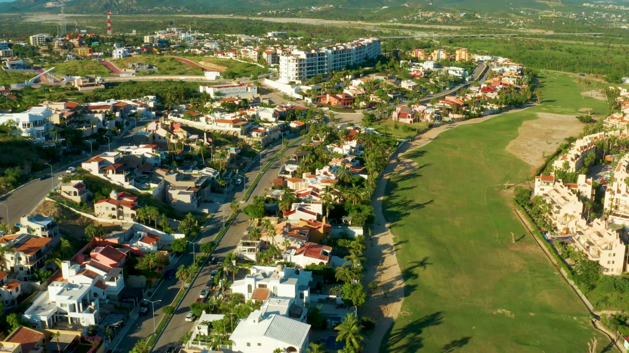 Opening aerial frame over San Jose Del Cabo homes, curving roads, palm trees, and a wide golf course fairway with no visible people.