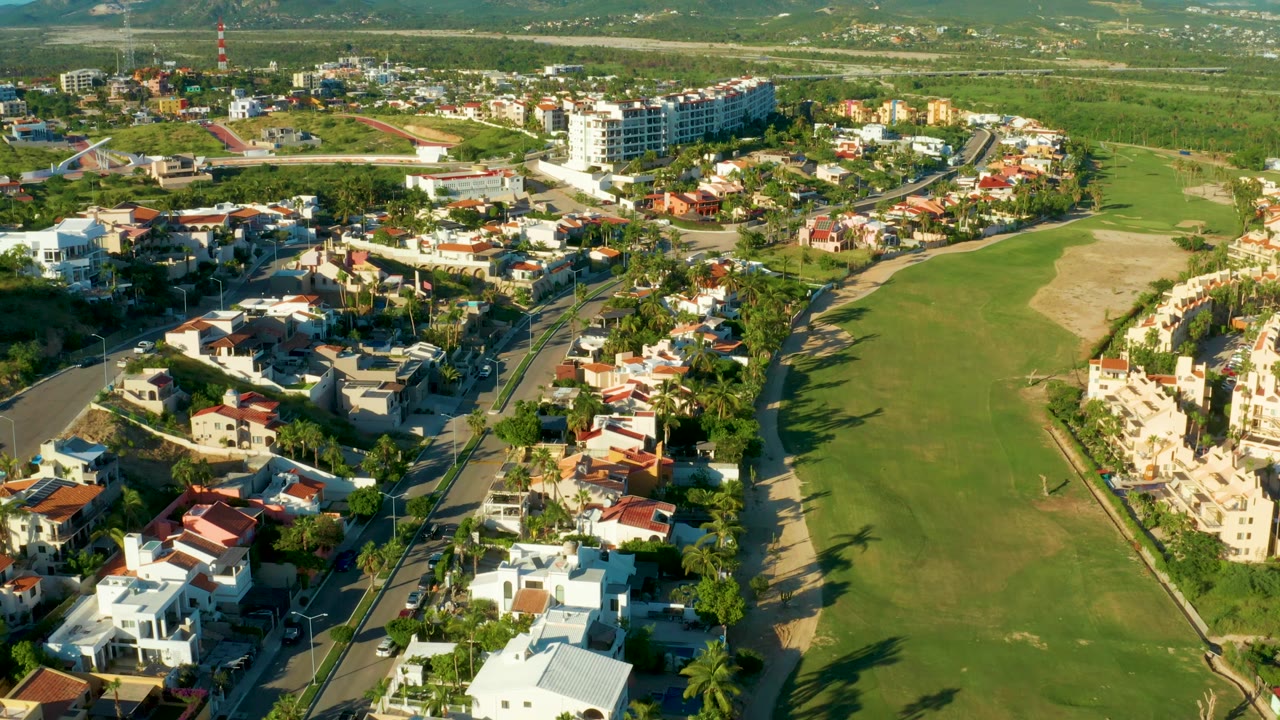 Aerial frame in San Jose Del Cabo showing more of the central fairway, clustered homes, roadside palms, and distant low hills with no visible people.