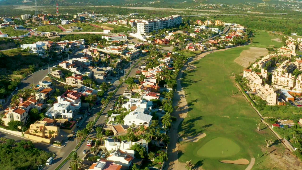 Aerial frame over San Jose Del Cabo showing white homes, terracotta roofs, a green fairway, curving residential roads, long shadows, and no visible people.