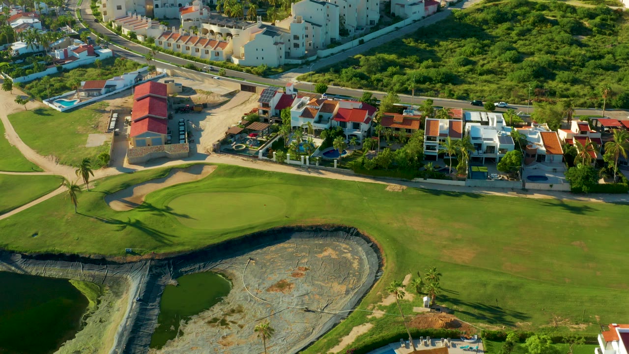 Aerial frame over San Jose Del Cabo showing a golf green and dark water hazard in the foreground, red-roofed homes and white residential buildings beyond, and no visible people.