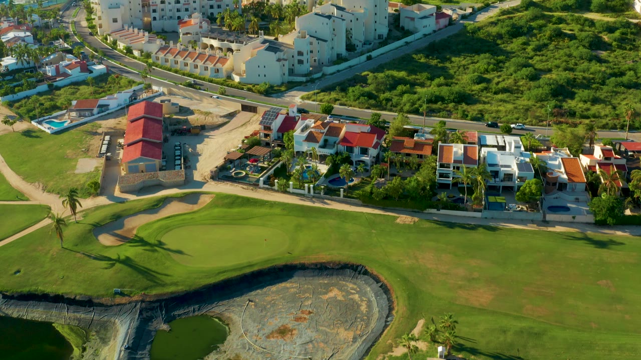 Drone frame from San Jose Del Cabo showing broad green course grass, a curved sand trap, neighboring homes with red and white roofs, a road, and no visible people.