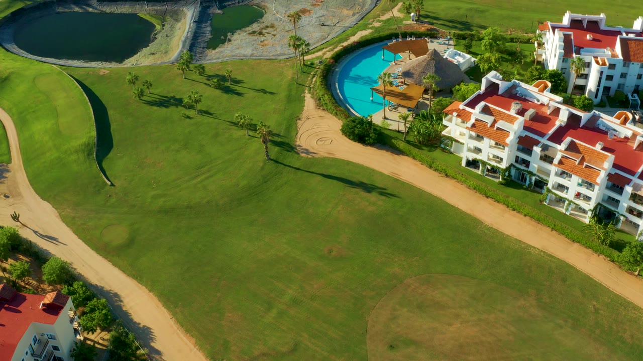 Aerial view in San Jose Del Cabo showing green fairway grass, two ponds, a bright blue pool, tan paths, and white red roof residences, with no visible people.