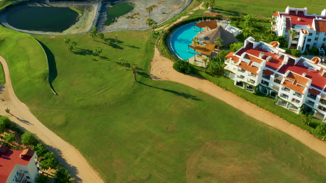 A high view in San Jose Del Cabo frames the fairway across the lower half, with dark ponds, a blue pool, red roof buildings, and curving tan paths, with no visible people.