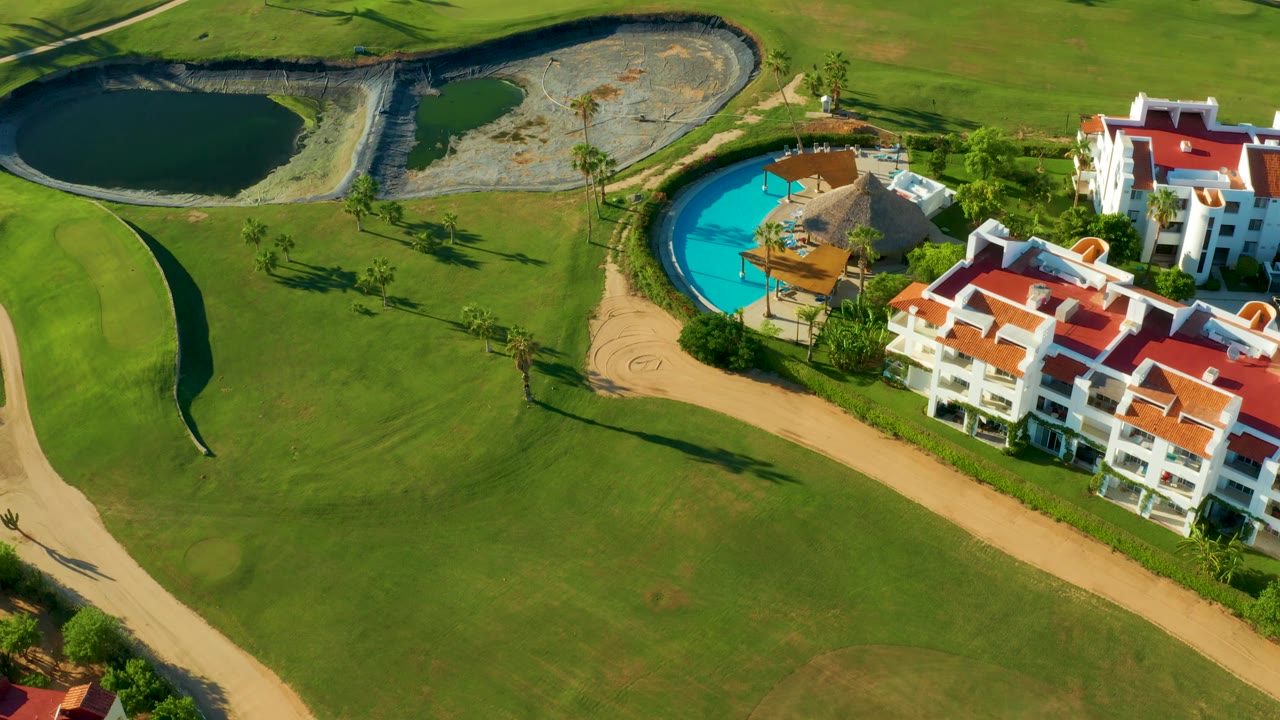 San Jose Del Cabo golf course housing appears from above, with red roof white buildings near a turquoise pool, dark water ponds, trimmed grass, and pathways, with no visible people.