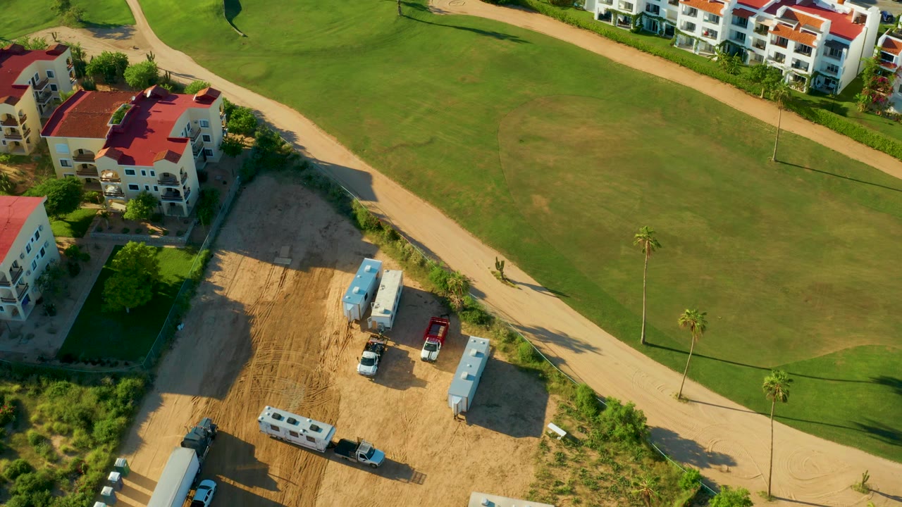 Aerial frame in San Jose del Cabo showing a construction lot with white trailers and vehicles beside a tan dirt road, green golf course grass, red-roofed residential buildings, and no visible people.