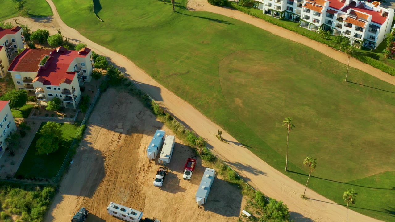 Aerial frame in San Jose del Cabo showing more open green fairway, the diagonal dirt road, red-roofed buildings along the top edge, part of the construction lot, and no visible people.