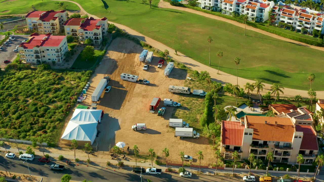 Aerial frame in San Jose Del Cabo showing a dirt construction lot with parked trailers and vehicles, red-roof residential buildings, a road at the bottom, a white tent roof, and no visible people.