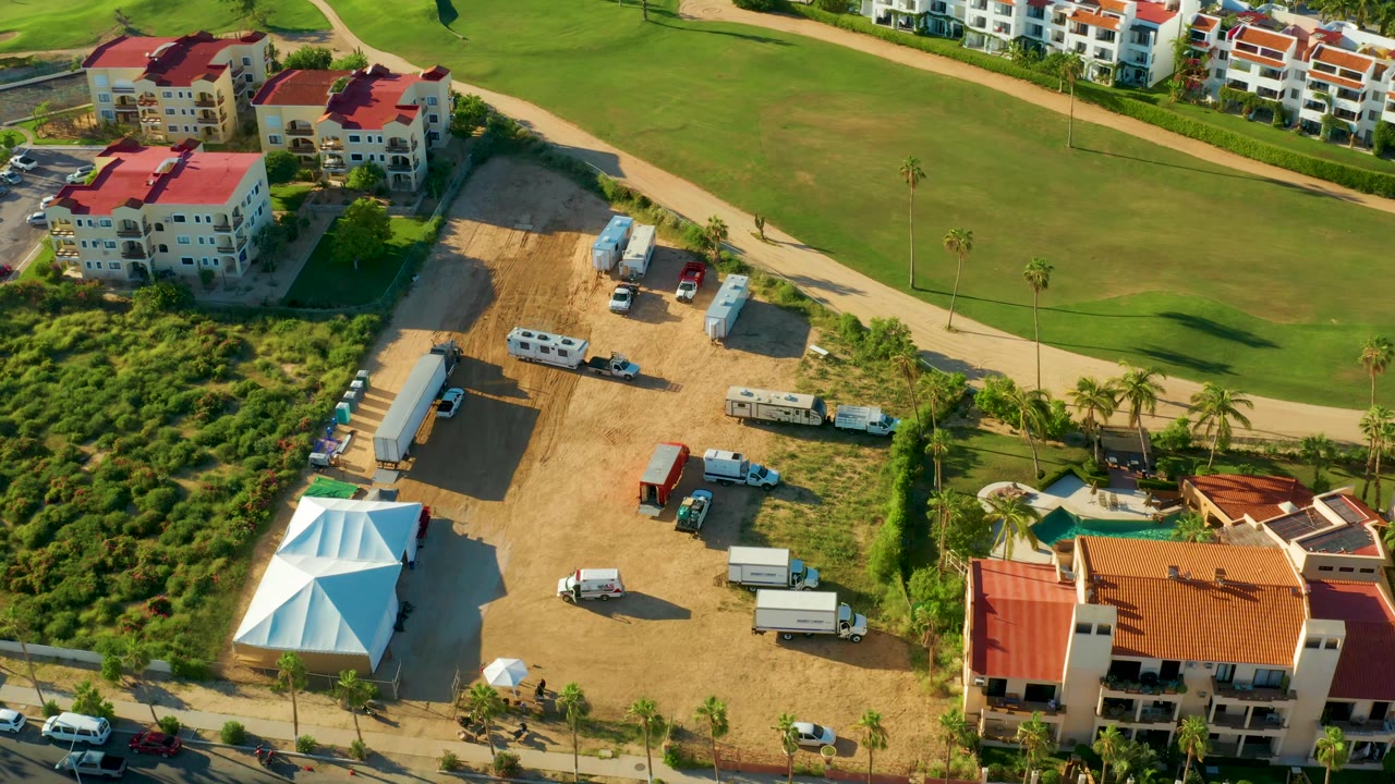Elevated San Jose Del Cabo view showing the dirt construction area and trailers centered between red-roof buildings, the road below, the green field above, and no visible people.