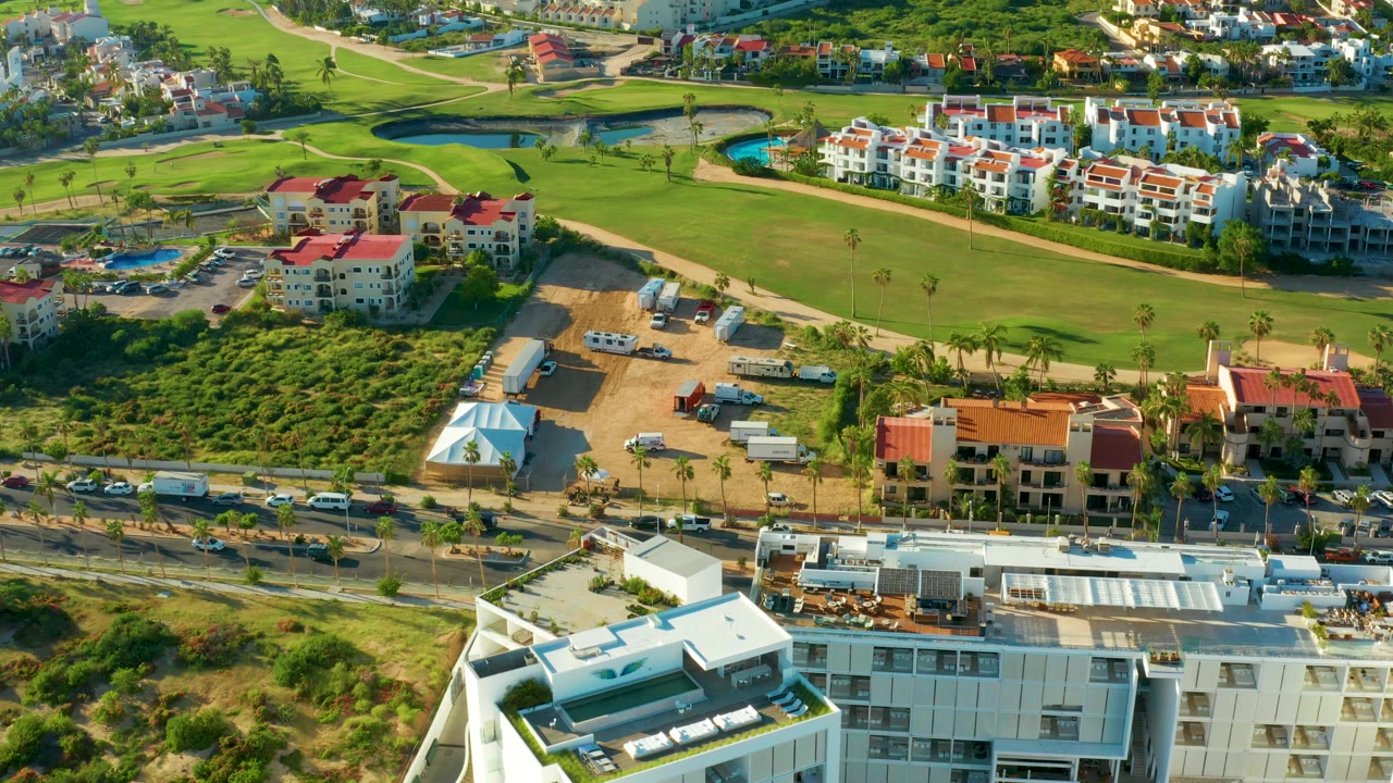 High aerial frame over San Jose del Cabo showing green fairways, a blue water hazard, red-roofed residential buildings, roads, construction lots, and no visible people.