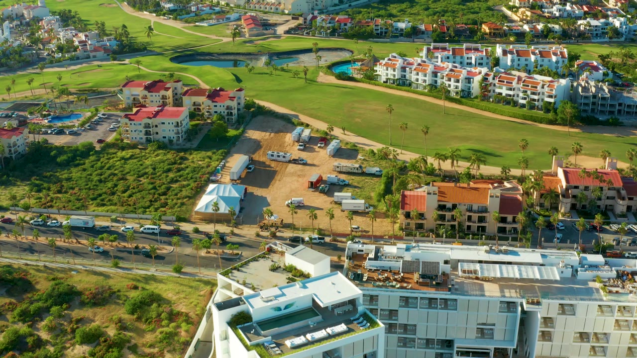 San Jose del Cabo aerial view with the fairway across the upper frame, white residential blocks and red roofs beyond it, work trucks near sandy lots, and no visible people.
