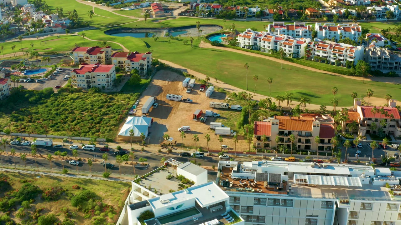 Drone frame in San Jose del Cabo showing the fairway sweeping behind clustered white buildings, a palm-lined road with cars, sandy construction ground, and no visible people.