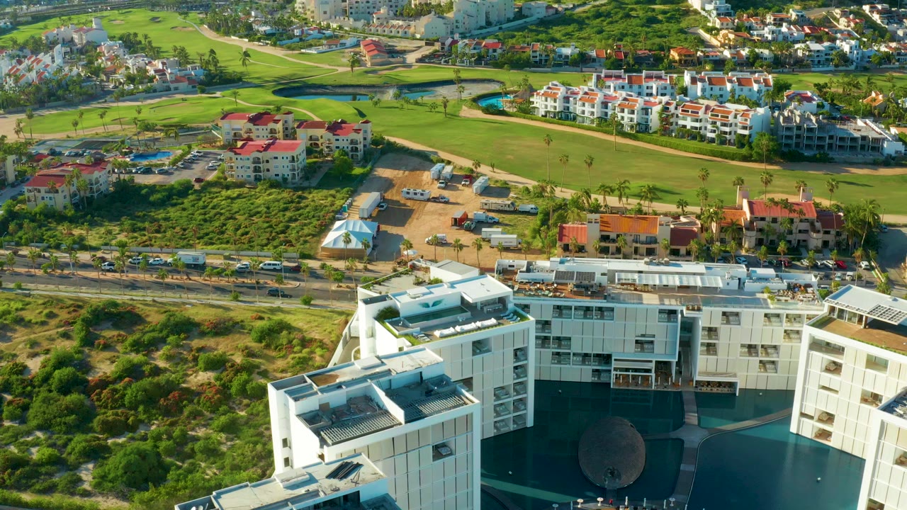 Aerial frame from San Jose Del Cabo, Mexico showing white multi-story buildings in the lower foreground, a road with cars, red-roof residences, green fields, pools, and no visible people.