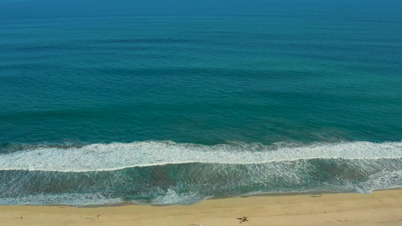 Overhead view of San Jose del Cabo surf, with deep blue water, a white foam line, tan sand, and no visible people at the start of the clip.