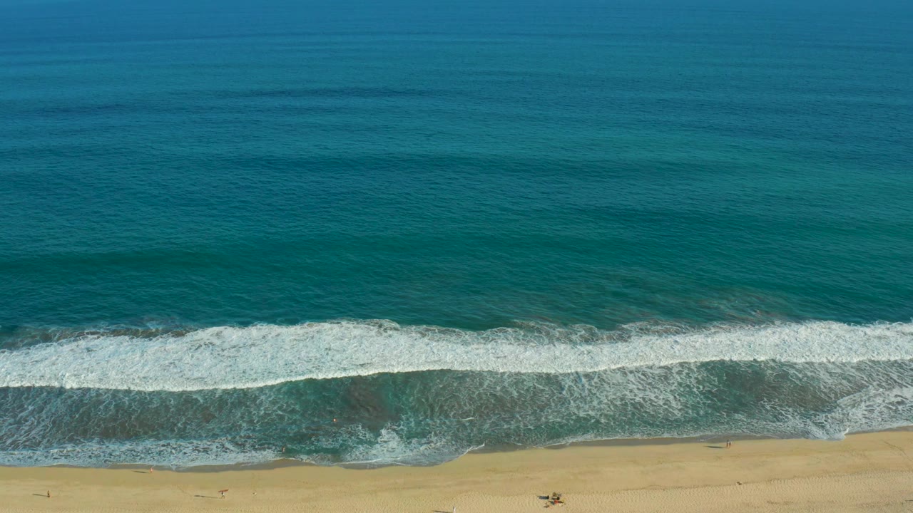 Aerial frame from San Jose del Cabo showing the foam band spread wider across shallow turquoise water before it reaches the sandy shore, with no visible people.