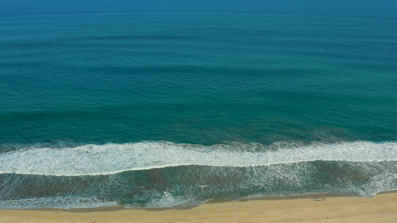 San Jose del Cabo shoreline seen from above, with a darker blue upper ocean, turquoise shallows, white surf, and a narrow tan beach with no visible people.