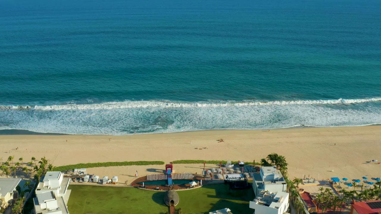 San Jose Del Cabo aerial view with teal ocean across the top, a continuous white wave band, open sand, and resort structures along the bottom edge, with no visible people.