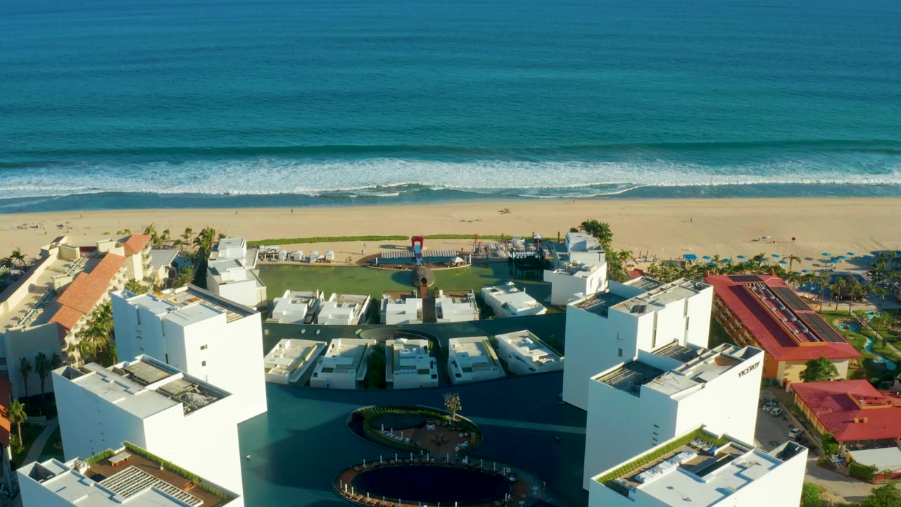 Opening aerial frame over San Jose Del Cabo, Baja California Sur, showing white resort buildings, red roof sections, a sandy beach, blue-green ocean, and no visible people.