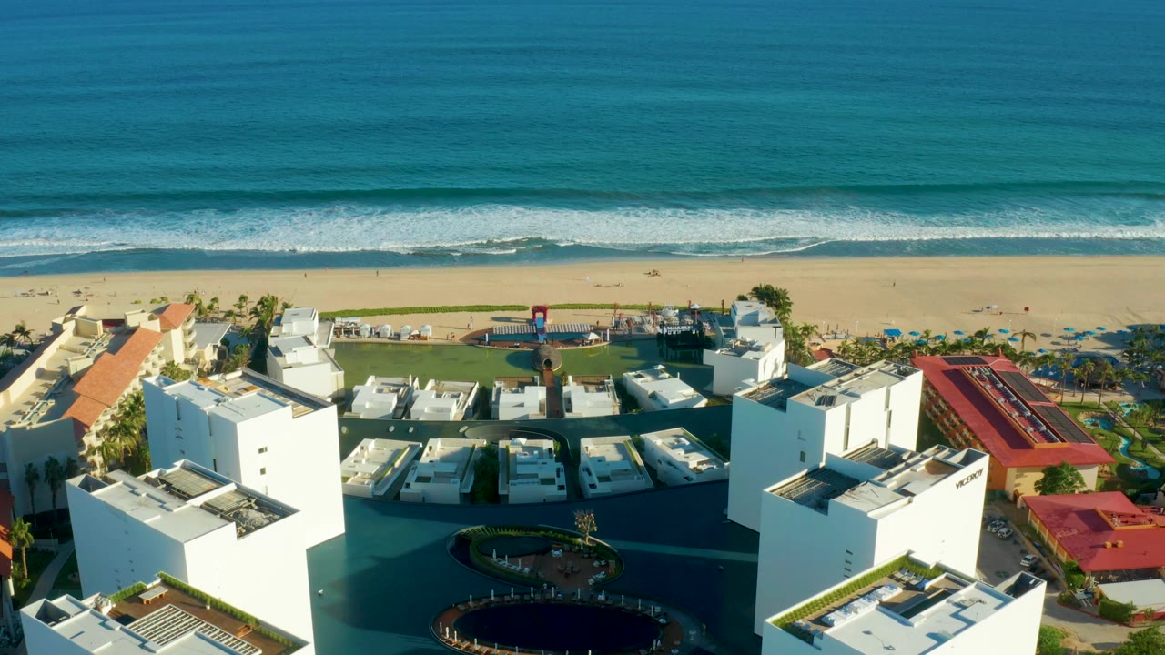 Aerial frame in San Jose Del Cabo with white flat-roof resort structures in the foreground, a narrow sandy beach band, rolling surf, and no visible people.