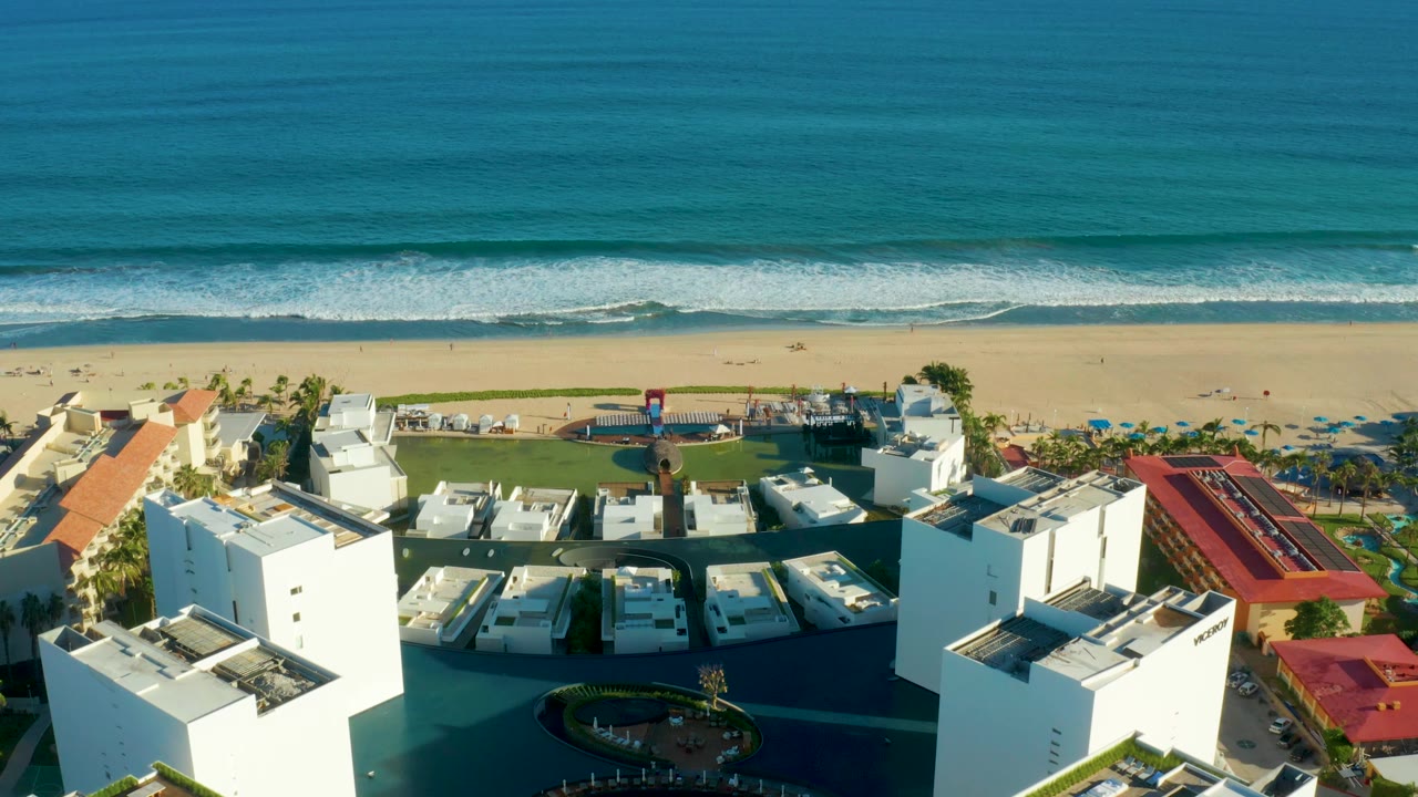 Aerial view over the San Jose Del Cabo resort grounds, with red roof strips, white buildings, beach umbrellas, sand, surf, blue water, and no visible people.