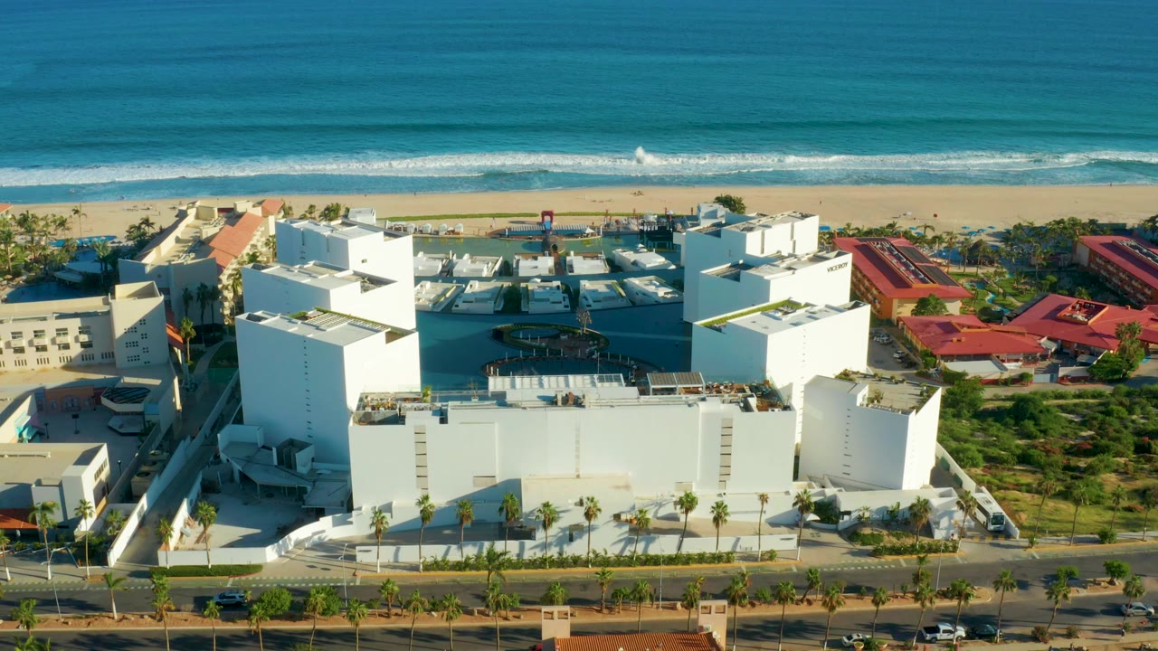 Aerial frame in San Jose del Cabo showing white hotel buildings, a beachfront road, sandy beach, and blue ocean with no visible people.