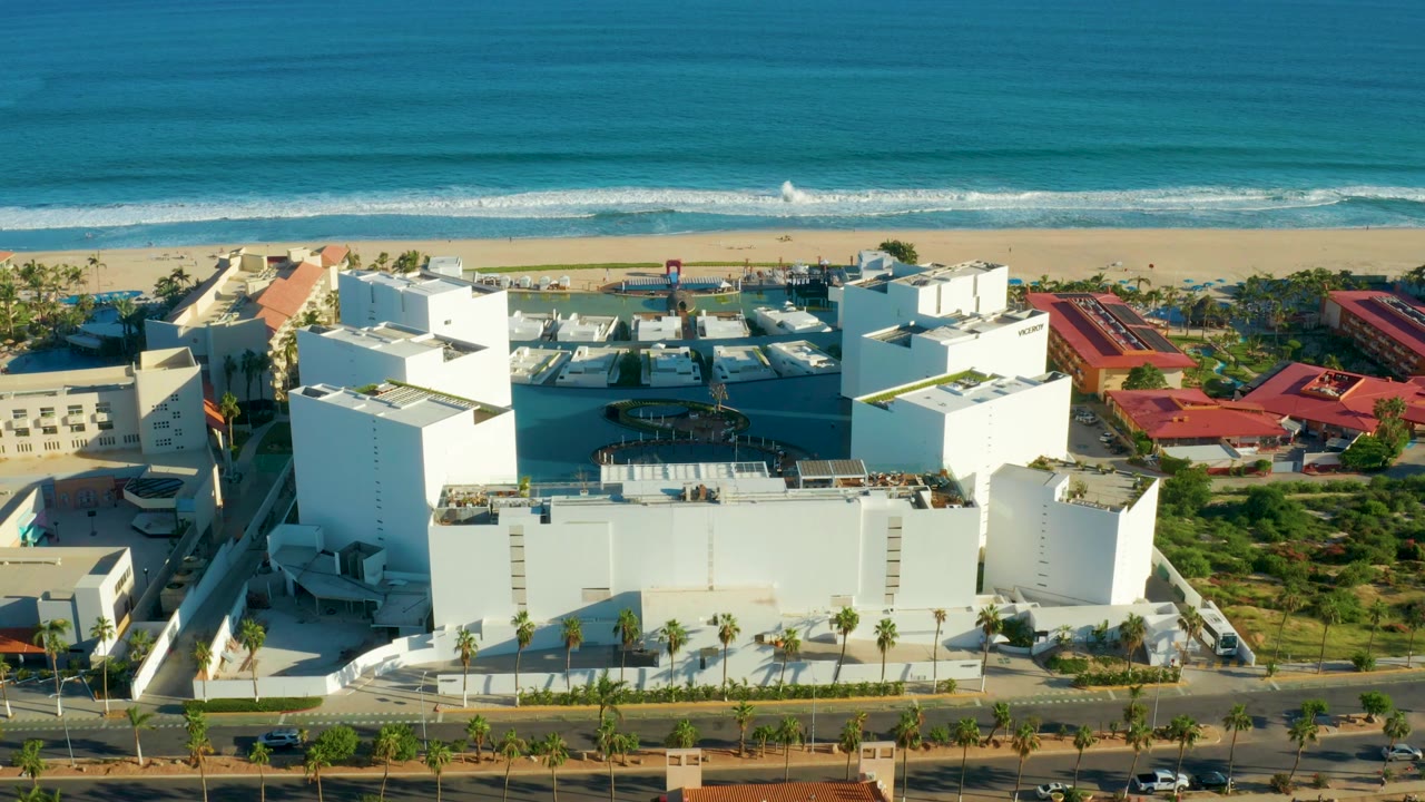 Aerial view in San Jose del Cabo with the white hotel complex more centered, a dark pool area visible, and surf lines behind the beach with no visible people.