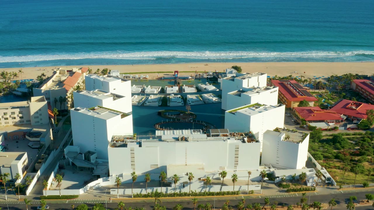 San Jose del Cabo aerial frame showing white hotel blocks, adjacent red-roofed buildings, palm-lined road, beach, and blue ocean with no visible people.