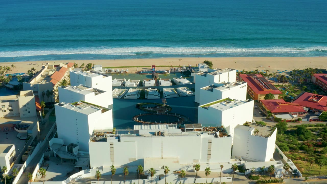 A high oblique frame in San Jose del Cabo looking over white hotel buildings toward a sandy beach, turquoise water, and a level ocean horizon with no visible people.