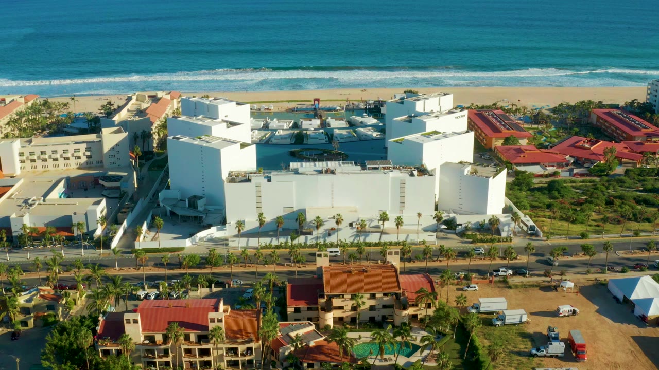 San Jose Del Cabo aerial view with white buildings centered below the beach, small vehicles on the road, surf lines offshore, and no visible people.
