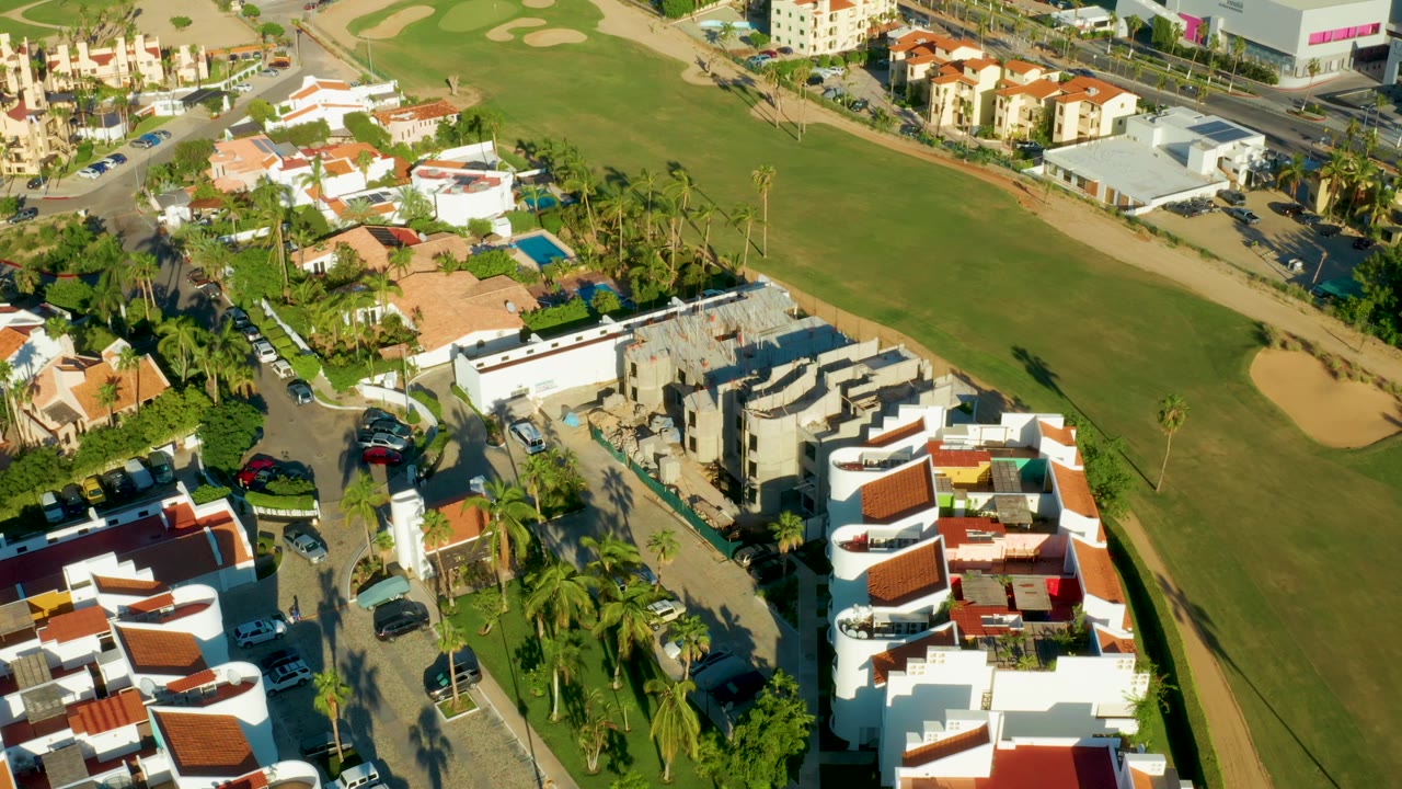 San Jose Del Cabo aerial frame with a cluster of white residences, red tile roofs, tall palms, parked vehicles, and the green baseball field running along the right side with no visible people.