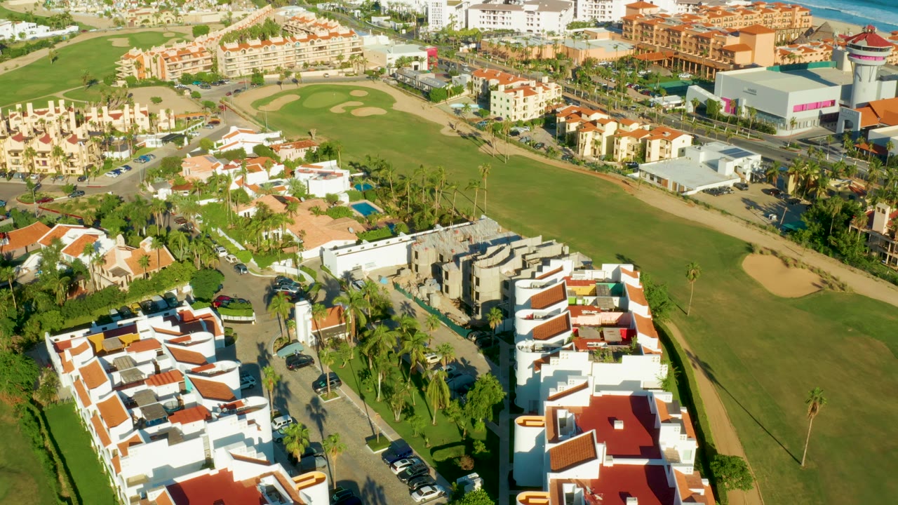 Aerial view in San Jose Del Cabo with the golf fairway running diagonally past white roofed homes, palms, parked cars, and surrounding low-rise development with no visible people.
