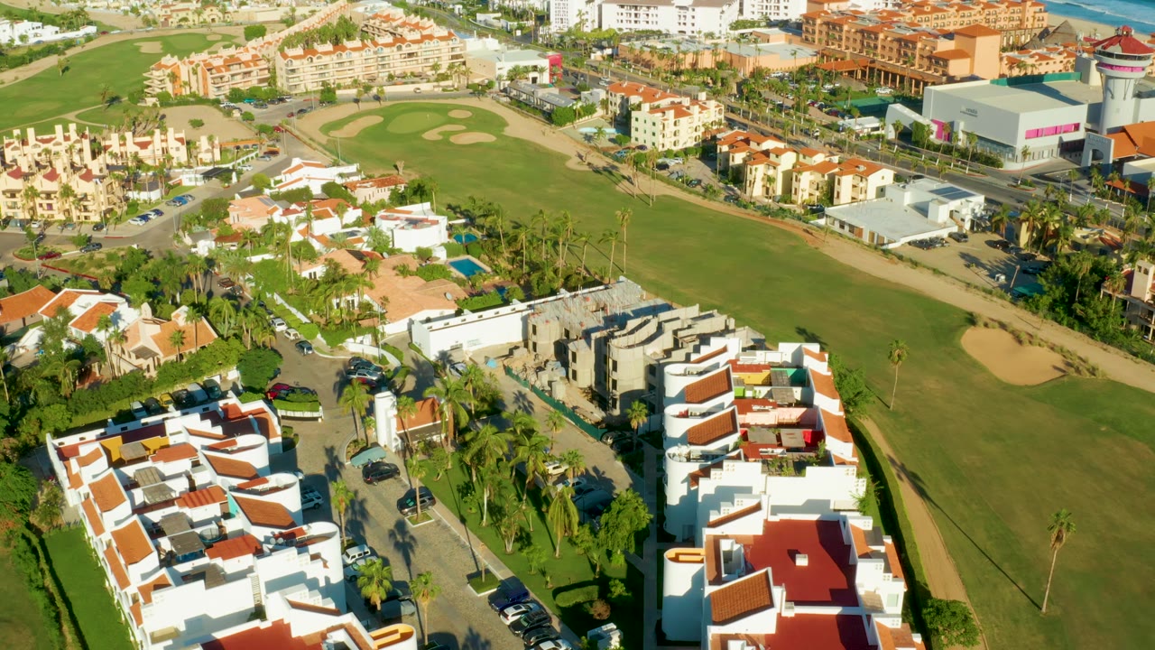 San Jose Del Cabo aerial frame showing white residential roofs in the foreground, cars along the road, palm landscaping, and golf greens and bunkers beyond, with no visible people.