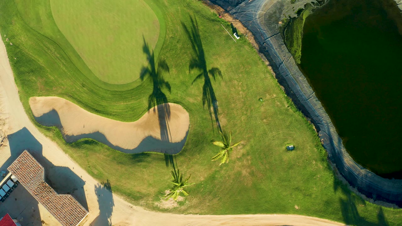 Top-down aerial frame in San Jose del Cabo showing a golf green, pale sand bunker, palm shadows, grass, a roof edge, and dark canal water, with no visible people.