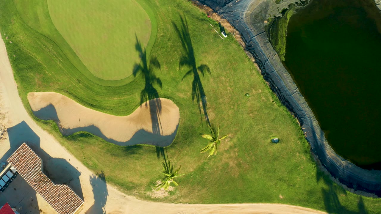 Overhead San Jose del Cabo view with the sand bunker and putting green on the left, long palm shadows across grass, and the dark waterway along the right edge, with no visible people.