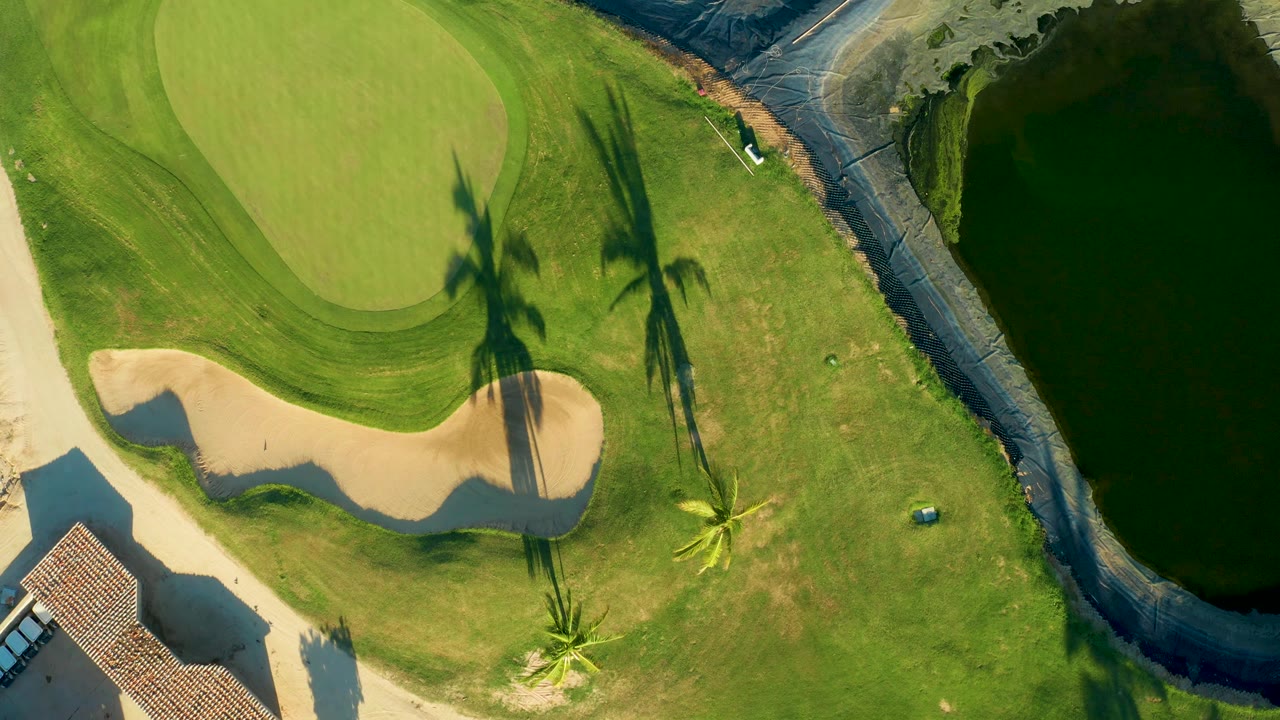 Top-down frame shows the San Jose del Cabo golf green and sand trap beside trimmed grass, palm shadows, a small blue course object, and dark canal water, with no visible people.